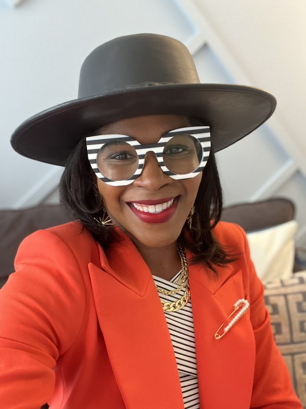 Heather Dowdy smiling in an orange blazer, a wide-brimmed dark hat and black-and-white striped glasses
