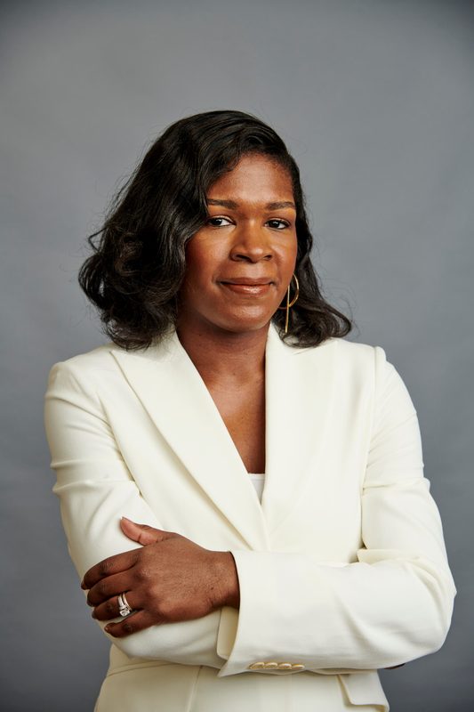 Heather Dowdy smiling in a white blazer, photographed against a gray studio backdrop