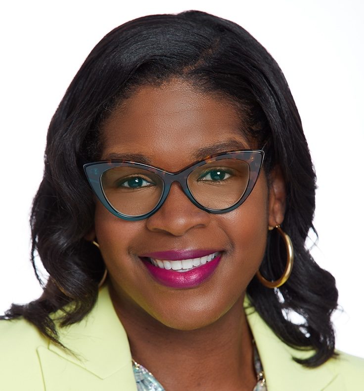 Heather Dowdy smiling in a yellow blazer, wearing dark cat-eye glasses and gold hoop earrings