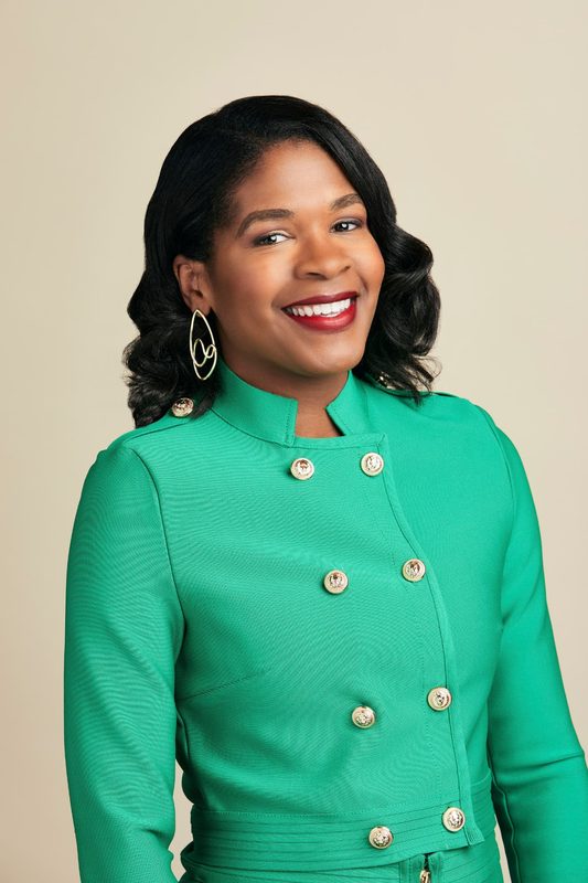 Heather Dowdy smiling in a green jacket with drop earrings against a soft beige background